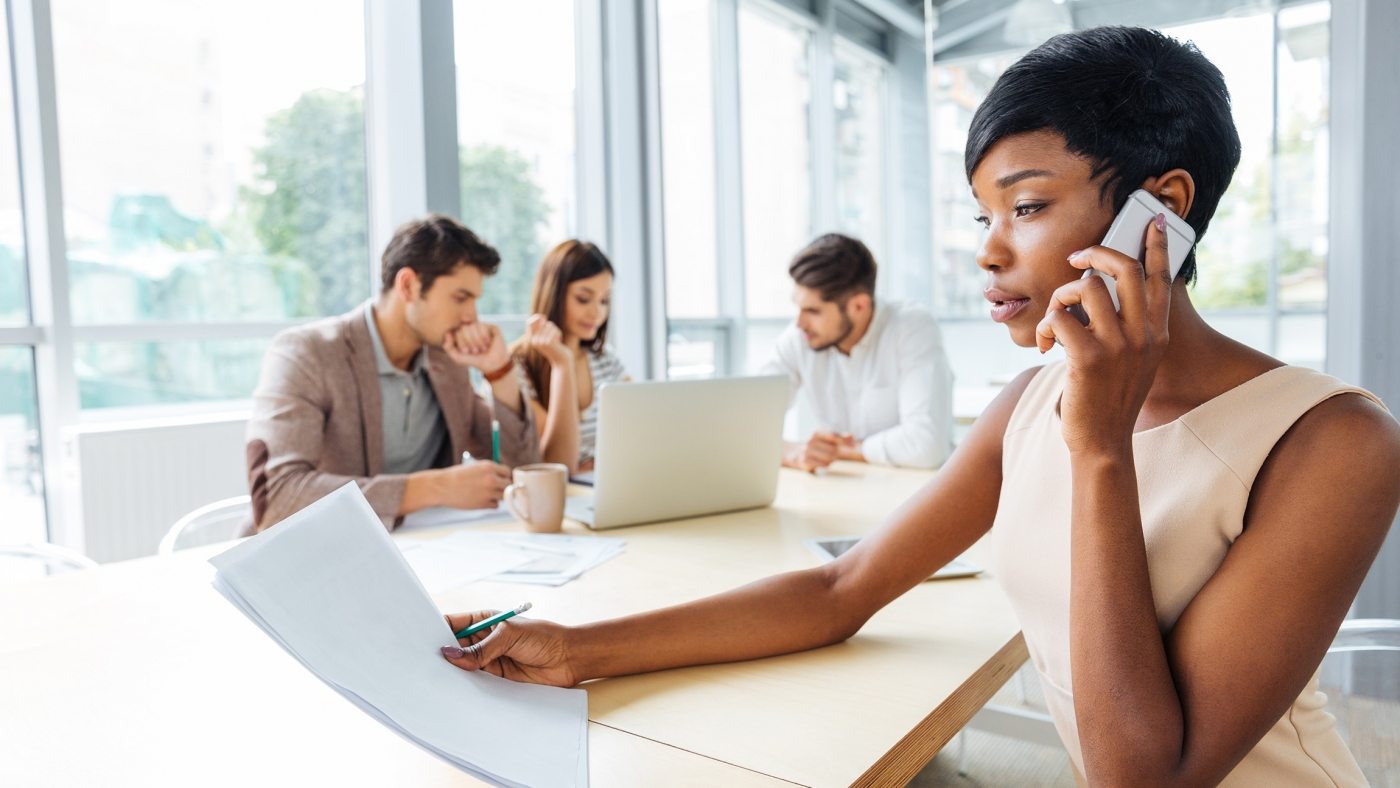 Serious successful young businesswoman with documents sitting and talking on cell phone in office